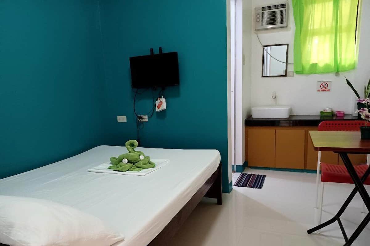 A small room with a bed, a desk, a chair, a TV mounted on the wall, and a bathroom visible through an open door.