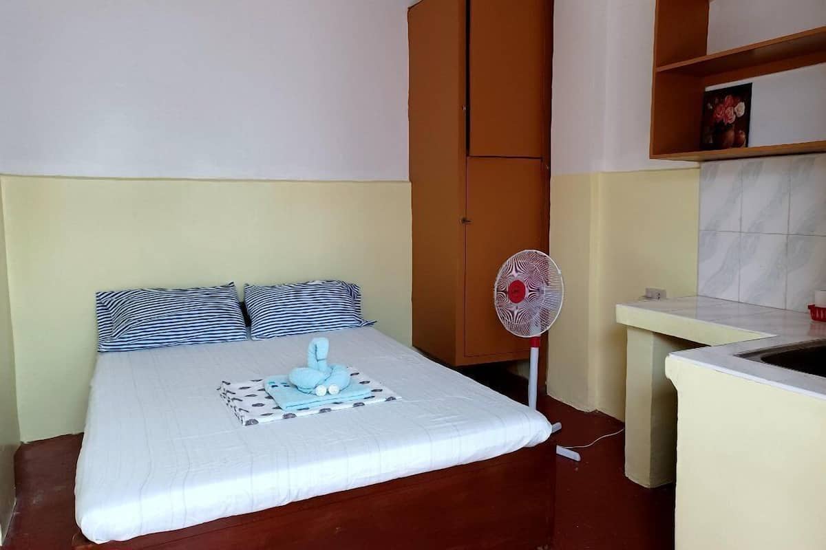 A small, compact room with a bed, a wardrobe, a fan, and a kitchenette.