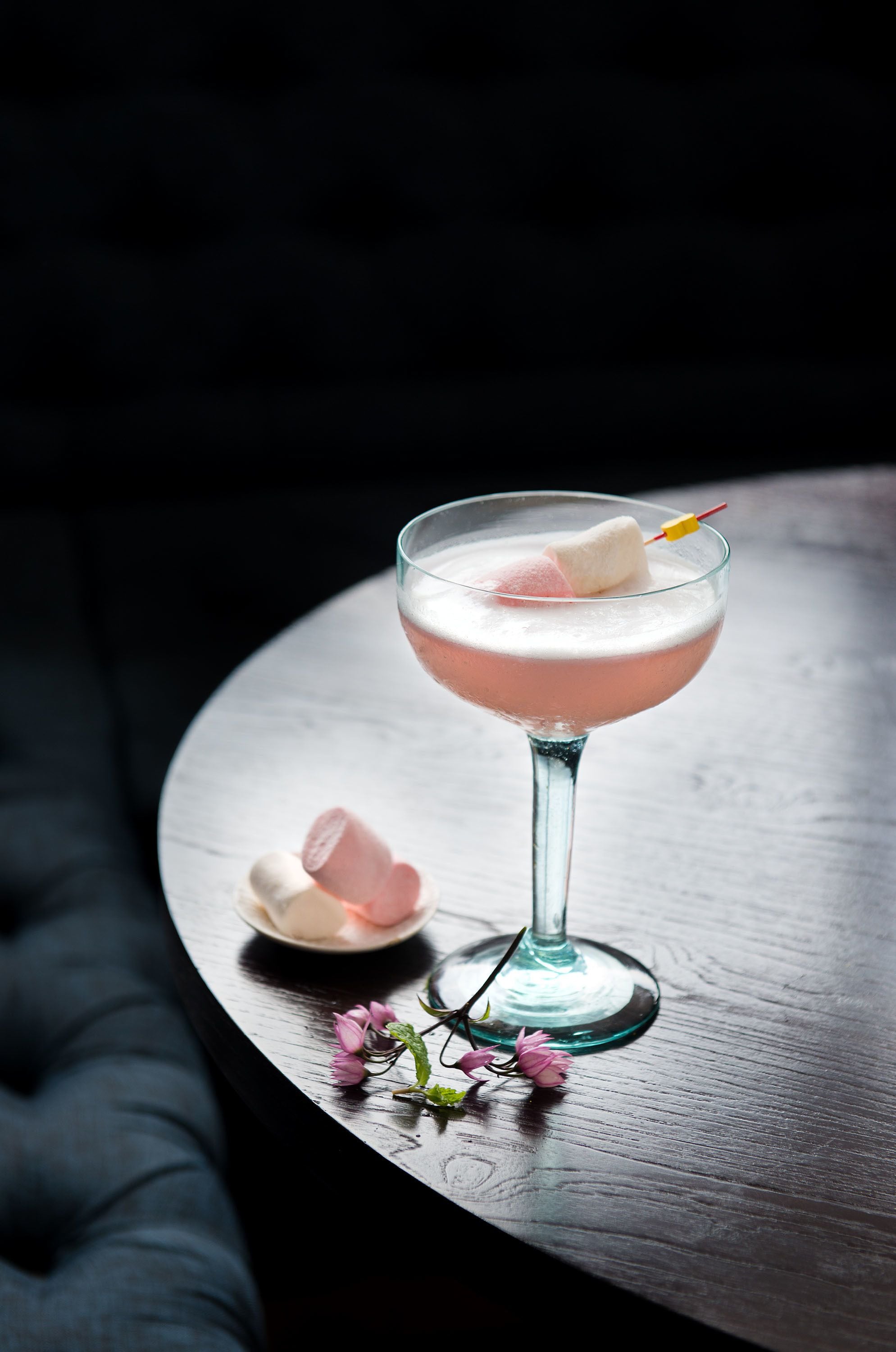 Un cóctel en un vaso con pajita amarilla, rodeado de flores rosadas y malvaviscos sobre una mesa de madera.