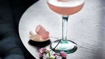Un cóctel en un vaso con pajita amarilla, rodeado de flores rosadas y malvaviscos sobre una mesa de madera.