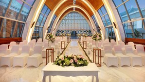 Un salón espacioso con techo abovedado alto y ventanas amplias, con filas de sillas blancas orientadas hacia una mesa central decorada con flores.