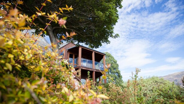 A wooden cabin with a balcony, surrounded by lush greenery and a clear sky. - Kauri House (Wanaka)