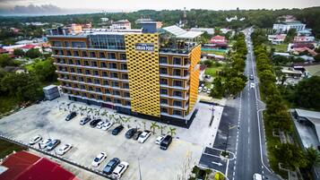 A multi-story hotel with a distinctive yellow and black facade, a parking lot with several cars, and a road with vehicles.