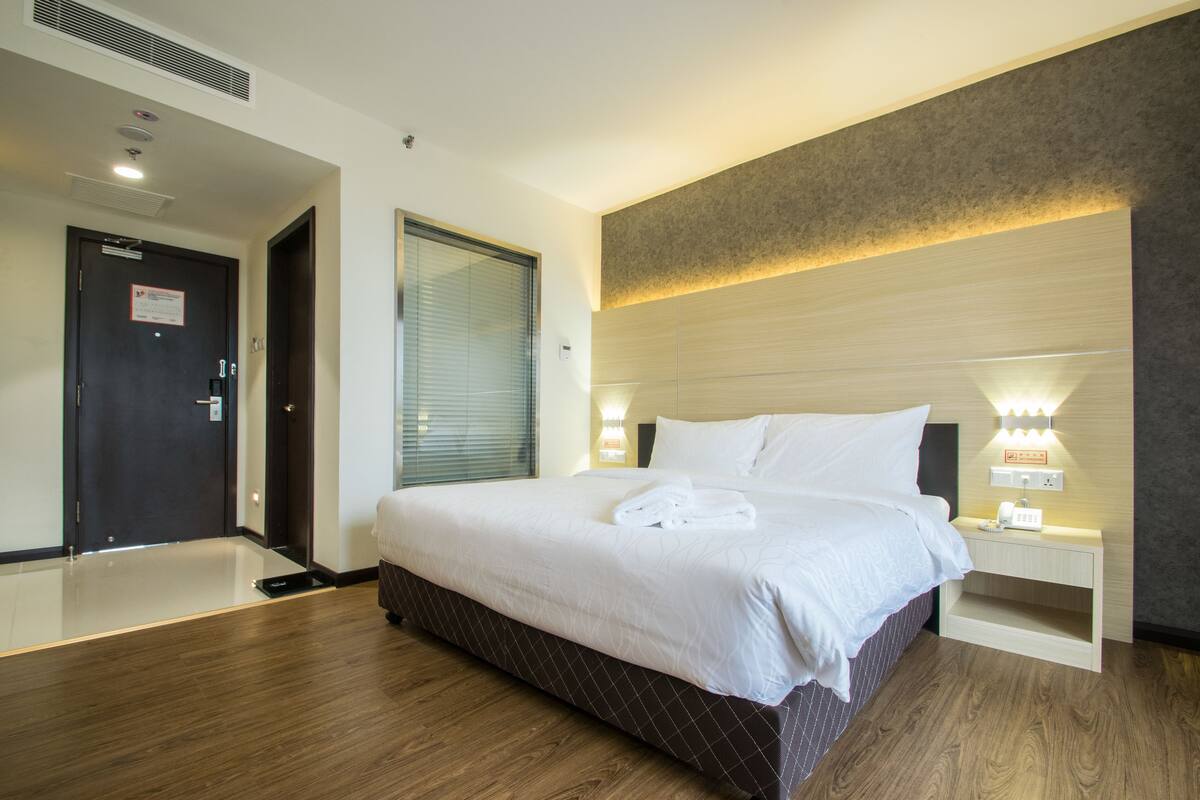 Quarto de hotel com uma cama grande, mesas de cabeceira, um criado-mudo, uma janela com persianas e uma porta.