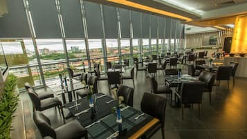 A modern restaurant with large windows offering city views, multiple dining tables with chairs, and a contemporary interior design.