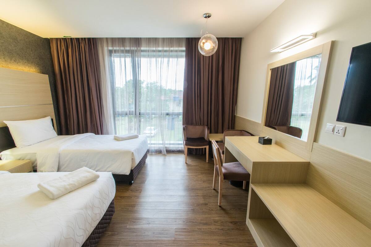 Quarto de hotel com duas camas, uma escrivaninha, uma cadeira, televisão e uma janela grande com cortinas.