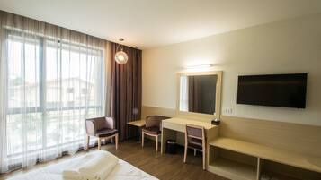 Habitación de hotel con cama, escritorio, silla, televisor y ventana grande con cortinas.