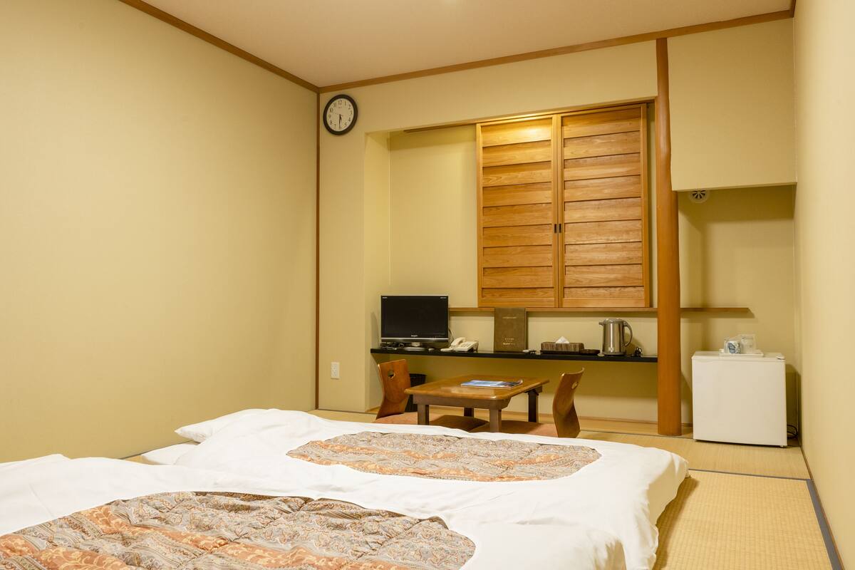 Una habitación de hotel con una cama, un escritorio con computadora, un refrigerador pequeño y un reloj en la pared.