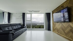 A modern living room with a black leather sofa, a large TV mounted on a wooden wall, and a large window offering a city view.