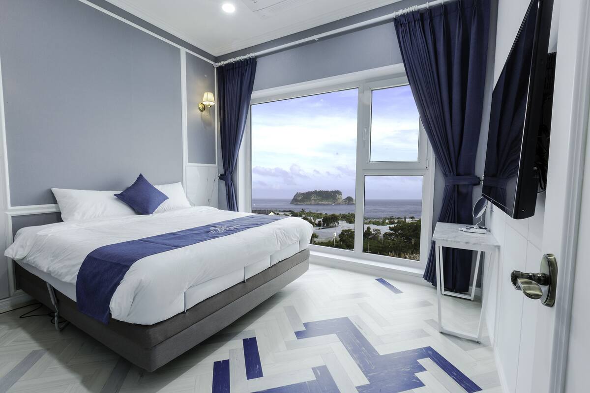 A modern hotel room with a large bed, a view of the sea, and a small table by the window.