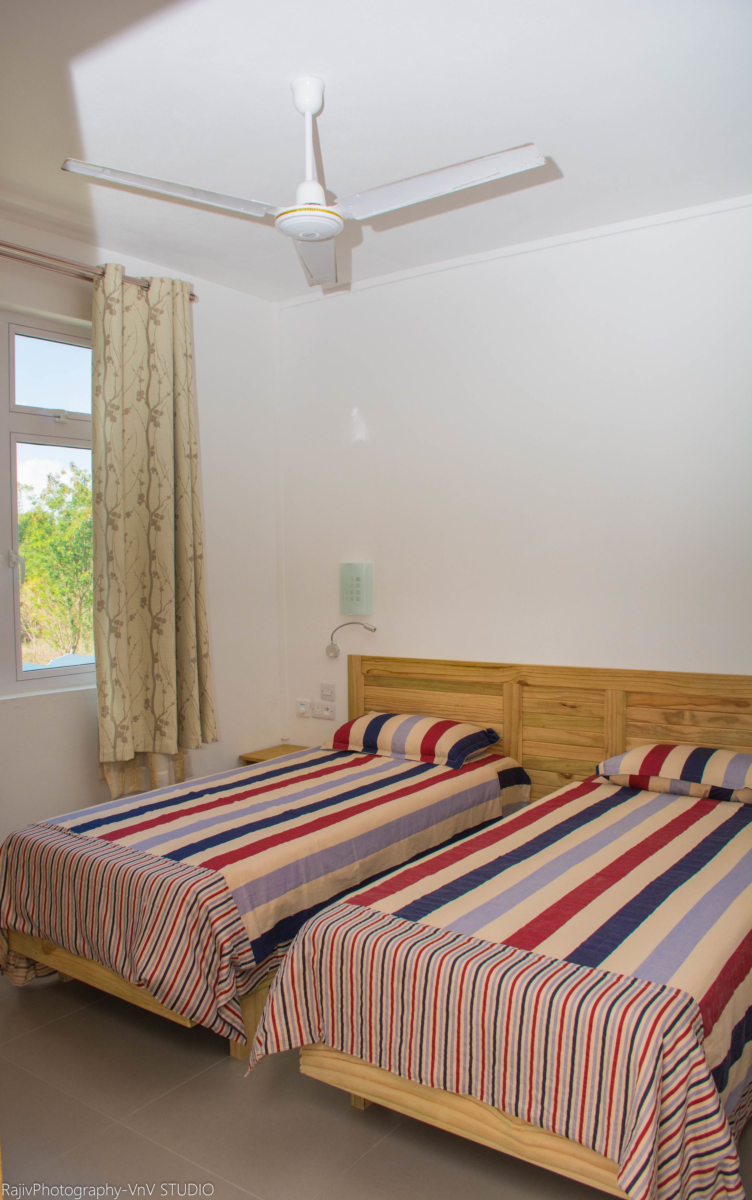 A bedroom with two single beds, a ceiling fan, a window with curtains, and a wall-mounted light switch.