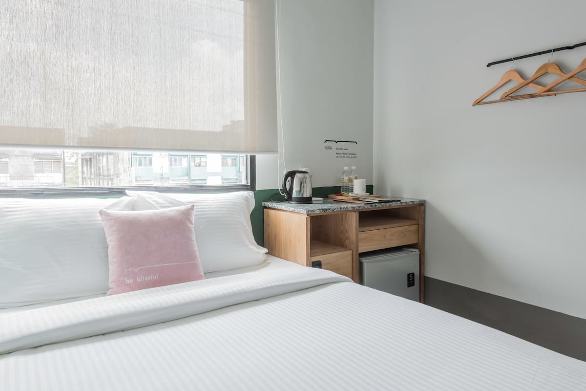 Habitación de hotel con cama, un pequeño escritorio con hervidor eléctrico, un refrigerador pequeño y ventana con vistas a edificios.