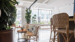 Un espace intérieur vaste et lumineux, aménagé avec des meubles en osier, des plantes en pot et de grandes fenêtres qui offrent une vue sur l’extérieur.