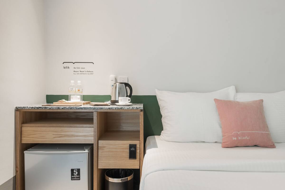 Habitación de hotel con cama, mesita de noche, cafetera, refrigerador y cubo de basura.