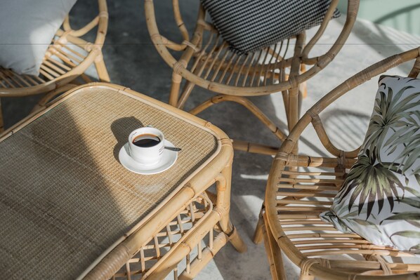 Wicker furniture with a woven table and chairs, a white cup with a saucer, and a patterned pillow.