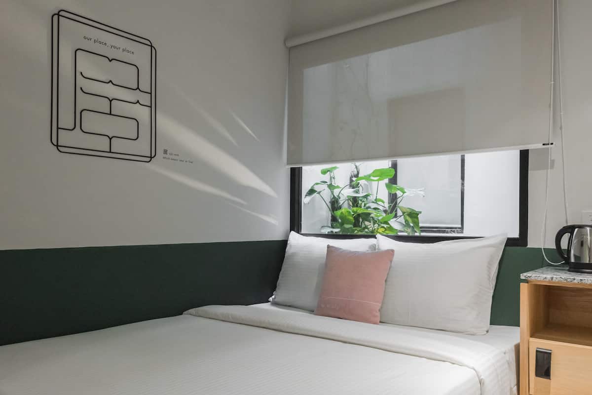 Una cama bien hecha con almohadas blancas y rosadas, un cabecero verde y una ventana con plantas.