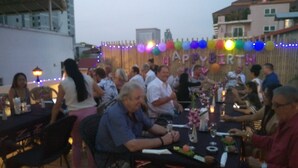 Un gruppo di persone sedute a tavoli all'aperto, con festoni e palloncini colorati sullo sfondo.