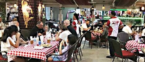 Un restaurante con manteles a cuadros rojos y blancos, clientes sentados en mesas y un camarero con uniforme rojo y blanco.