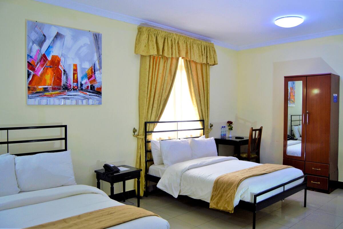 A hotel room with two beds, a desk, a chair, a wardrobe, and a painting on the wall.