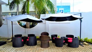 An outdoor seating area with wicker furniture and umbrellas, surrounded by a white wall and a palm tree. - M Hotel Mbezi (Dar es Salaam)