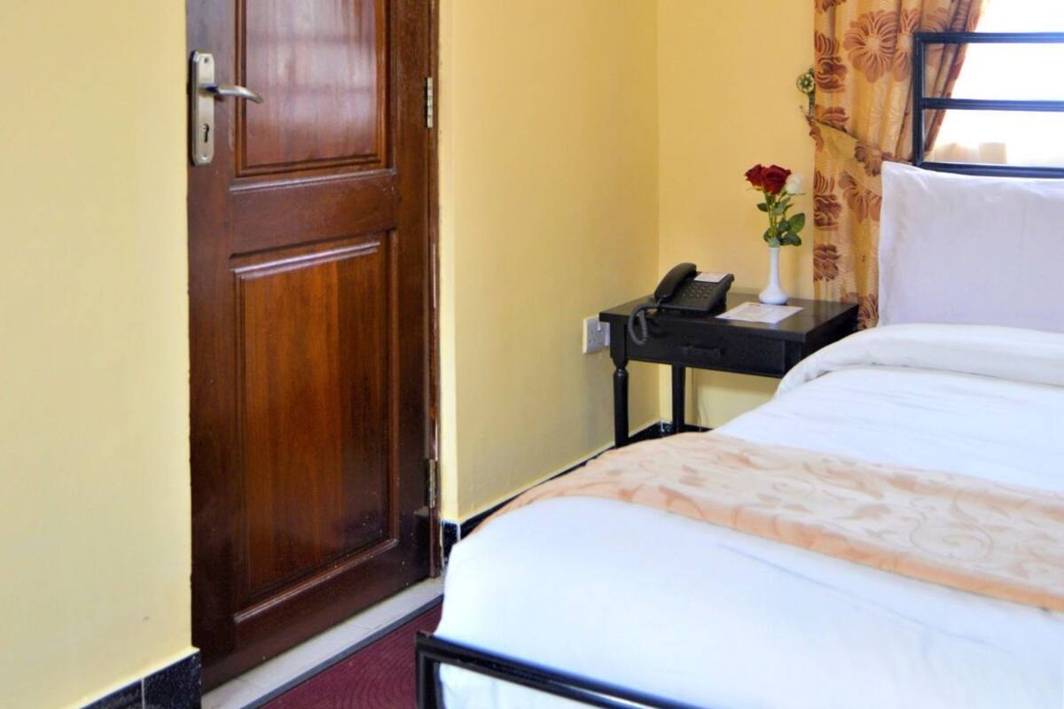 A hotel room with a wooden door, a bedside table with a phone and a vase of flowers, a bed with a patterned bedspread, and a window with floral curtains.