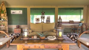 Um quarto acolhedor com sofá, duas cadeiras, uma mesa de madeira com objetos decorativos, uma estante e uma janela com cortinas verdes.