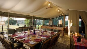 Un área de comedor cubierta con carpas, con una larga mesa dispuesta para una comida, rodeada de sillas.