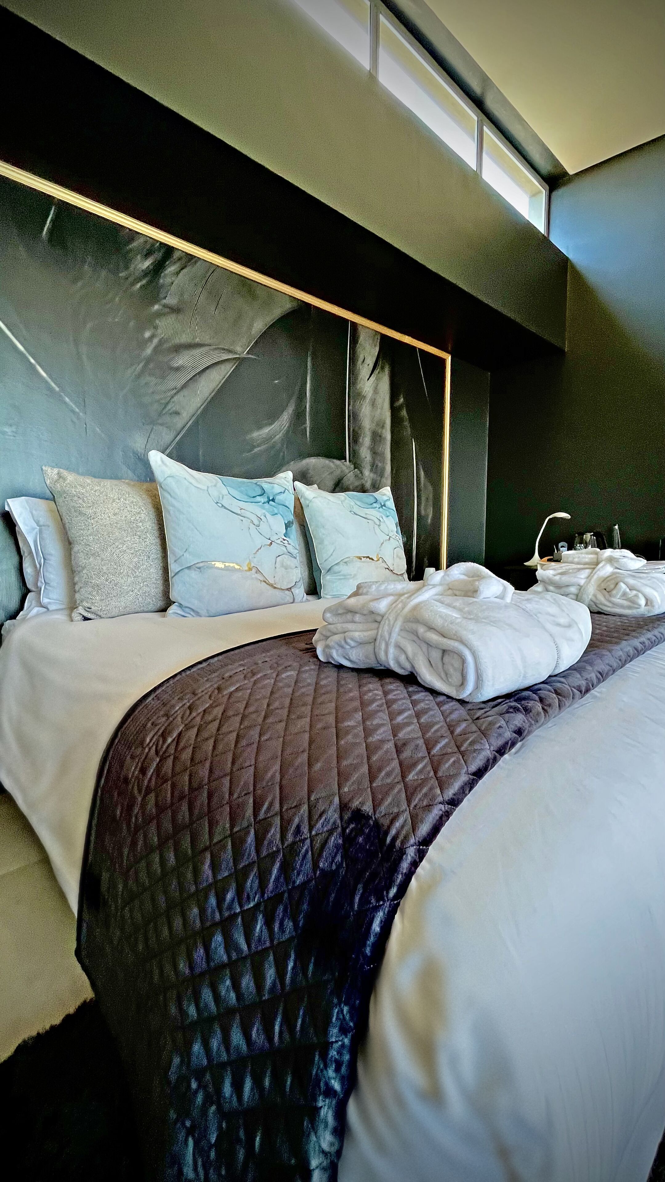 A modern hotel room with a large bed, a dark headboard, and a view of the sky through a skylight.