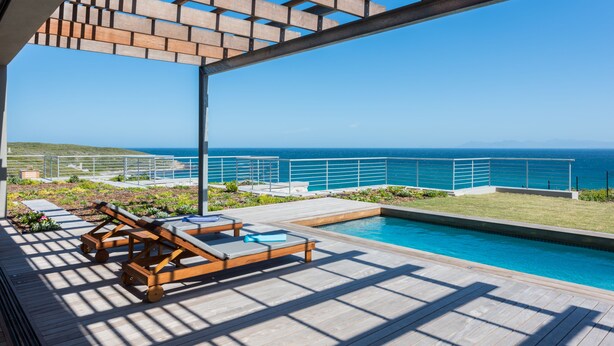 A poolside area with wooden loungers, a small pool, and a view of the ocean. - Romans Villa (Gansbaai)
