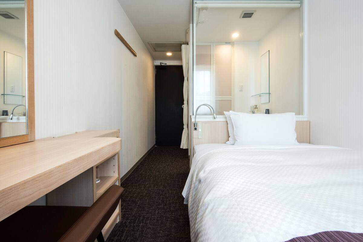 Une chambre d’hôtel avec un bureau en bois, un banc, un lit avec une literie blanche, un miroir et une fenêtre avec des rideaux.