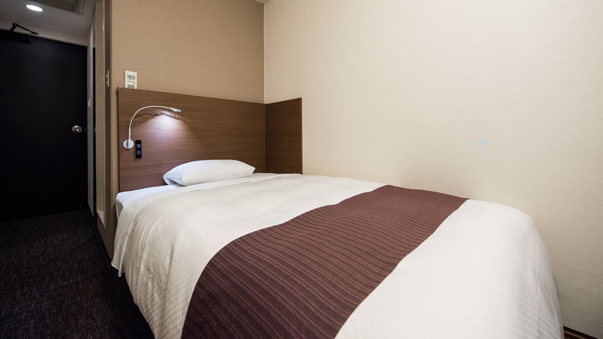 Une chambre d’hôtel avec un lit, une tête de lit, une lampe fixée au mur et une porte.