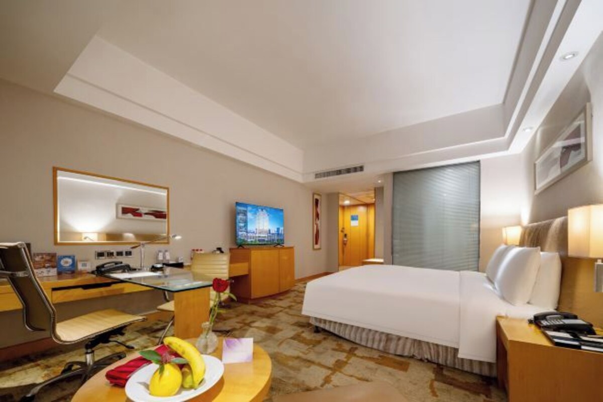 Habitación de hotel con cama, escritorio con computadora, silla, mesita con fruta y televisor en la pared.