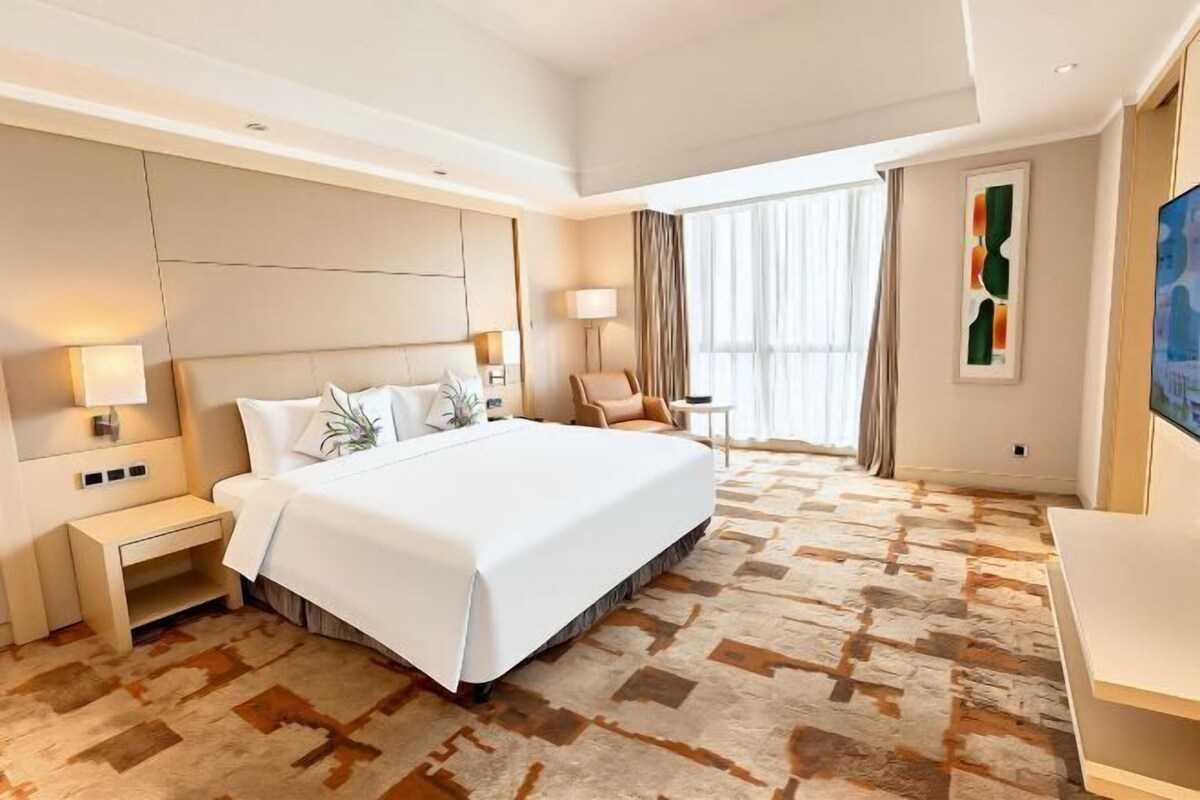 Ein Hotelzimmer mit einem großen Bett, Nachttischen, einem Sessel, einem Fernseher und einem Fenster mit Vorhängen.