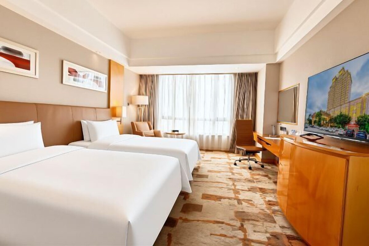 Ein Hotelzimmer mit zwei Betten, einem Schreibtisch, einem Fernseher und einem großen Fenster mit Vorhängen.