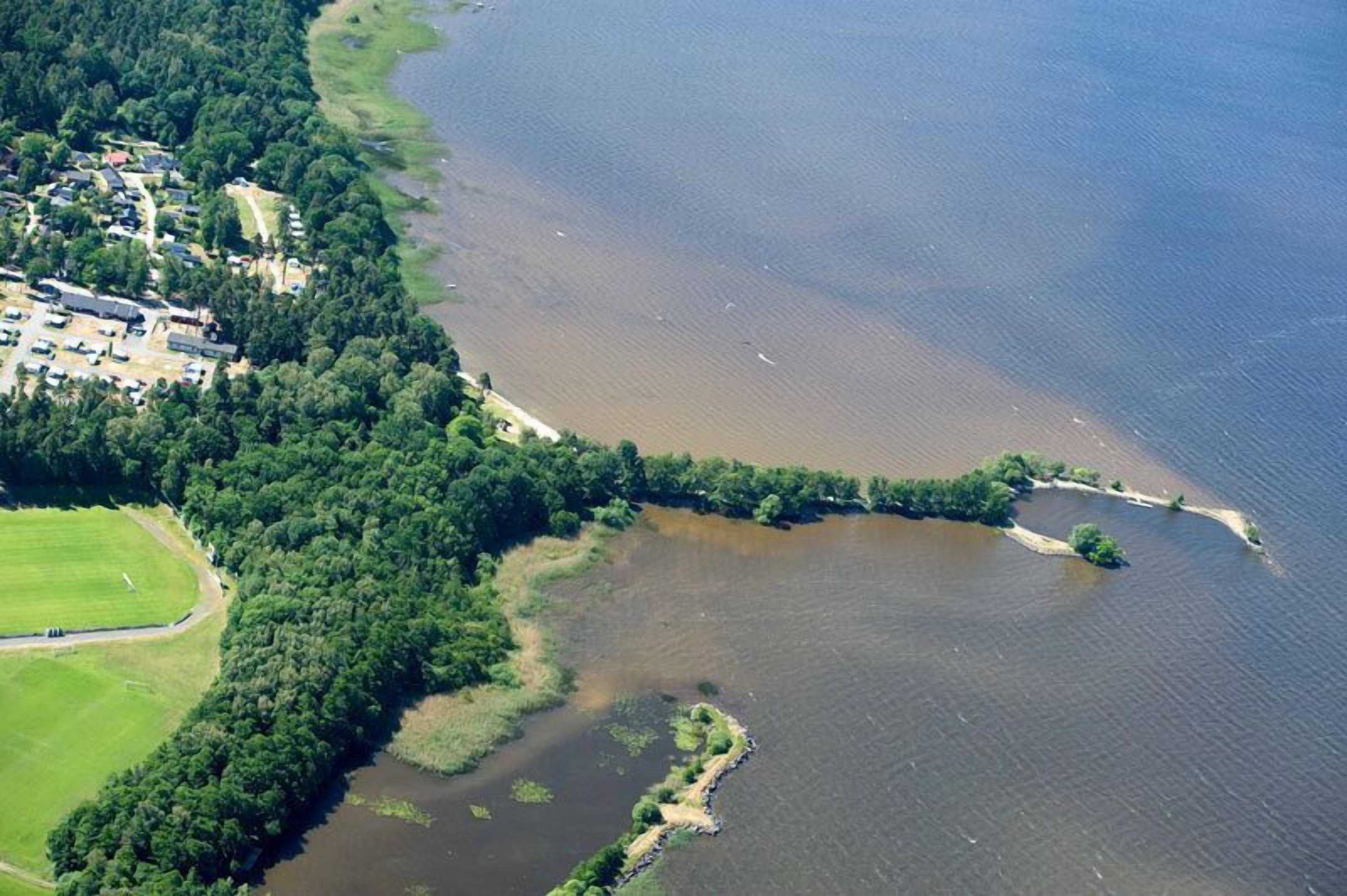 Vista aérea de una zona costera con un bosque denso, una pequeña isla y una área urbanizada con edificios y una carretera.