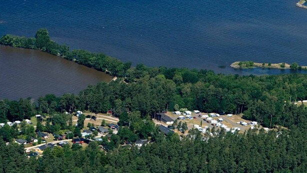 An aerial view of a lakeside campground with RVs and cabins, surrounded by dense forest and a dock extending into the lake.