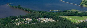 Vista aérea de un camping junto al lago con vehículos recreativos y cabañas, rodeado de un bosque denso y un muelle que se adentra en el lago.