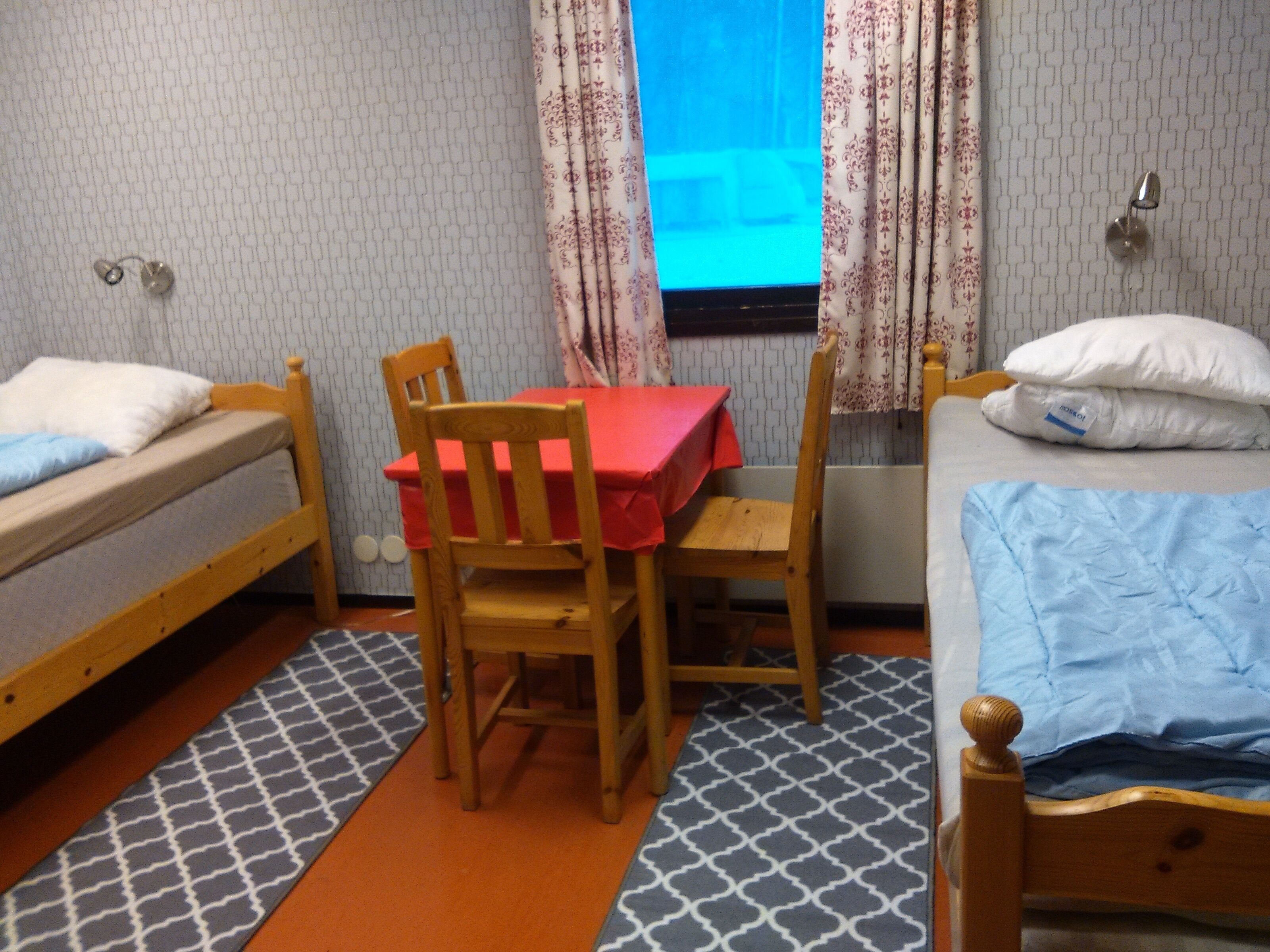 Een kamer met twee eenpersoonsbedden, een houten tafel met een rode tafelkleed en een stoel.