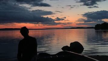 Una persona de pie junto a un cuerpo de agua tranquilo al atardecer, con un barco cerca.