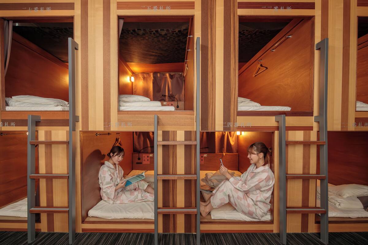 Une chambre d’hôtel avec des lits superposés, des murs en bois et une échelle. On y trouve deux lits avec des oreillers et des couvertures, et une personne est assise en train de lire un livre.