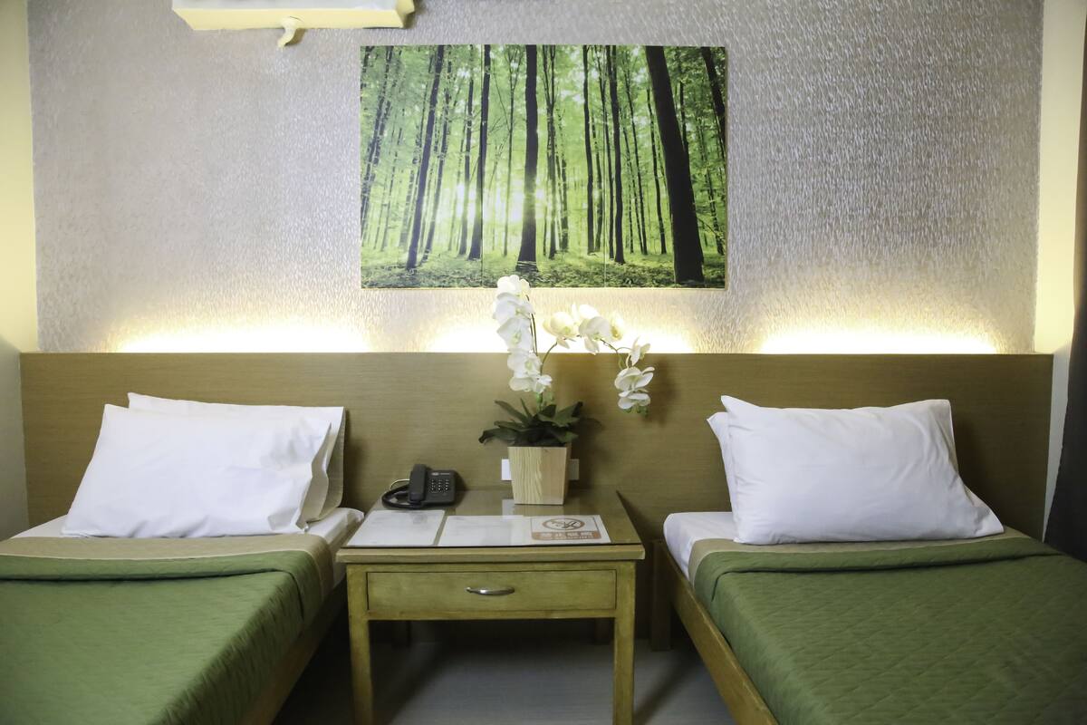Ein Hotelzimmer mit zwei Einzelbetten, einem kleinen Schreibtisch mit Telefon und Unterlagen, einem Nachttisch mit Lampe sowie einem an der Wand hängenden Bild von einem Wald.