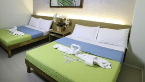 A hotel room with two single beds, each with a green quilt, white pillows, and neatly folded towels. There is a small wooden nightstand with a vase of white flowers between the beds. - Quoalla Hotel Makati (Makati)