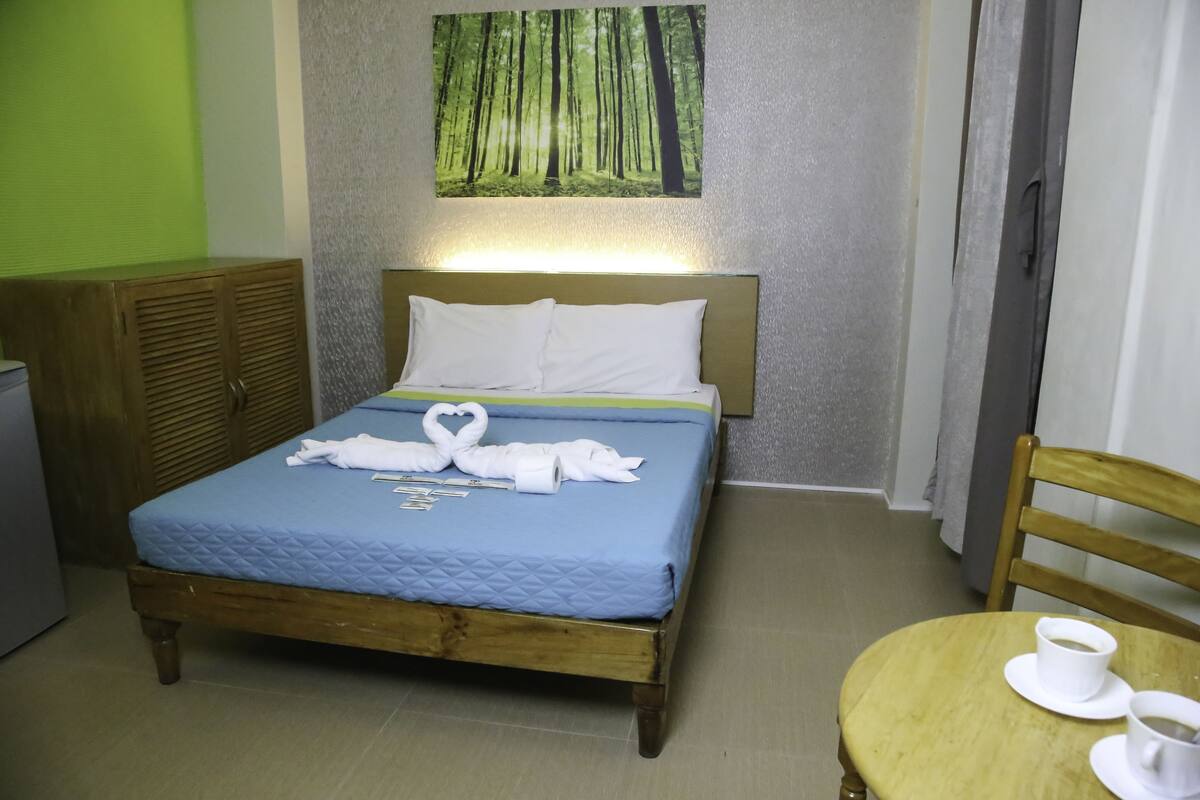 Ein ordentlich bezogenes Bett mit blauer Steudecke und weißen Kissen, ein hölzerner Kopfteil und ein Gemälde eines Waldes an der Wand.