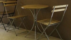 A small, round table with two metal-framed chairs. The chairs have wooden slats and metal legs. The setting appears to be indoors with a plain wall and tiled floor. - Quoalla Hotel Makati (Makati)