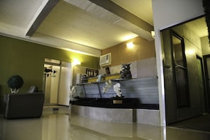 A reception area with a reception desk, a sofa, and a plant. There is a door leading to another room and an air conditioning unit on the wall.