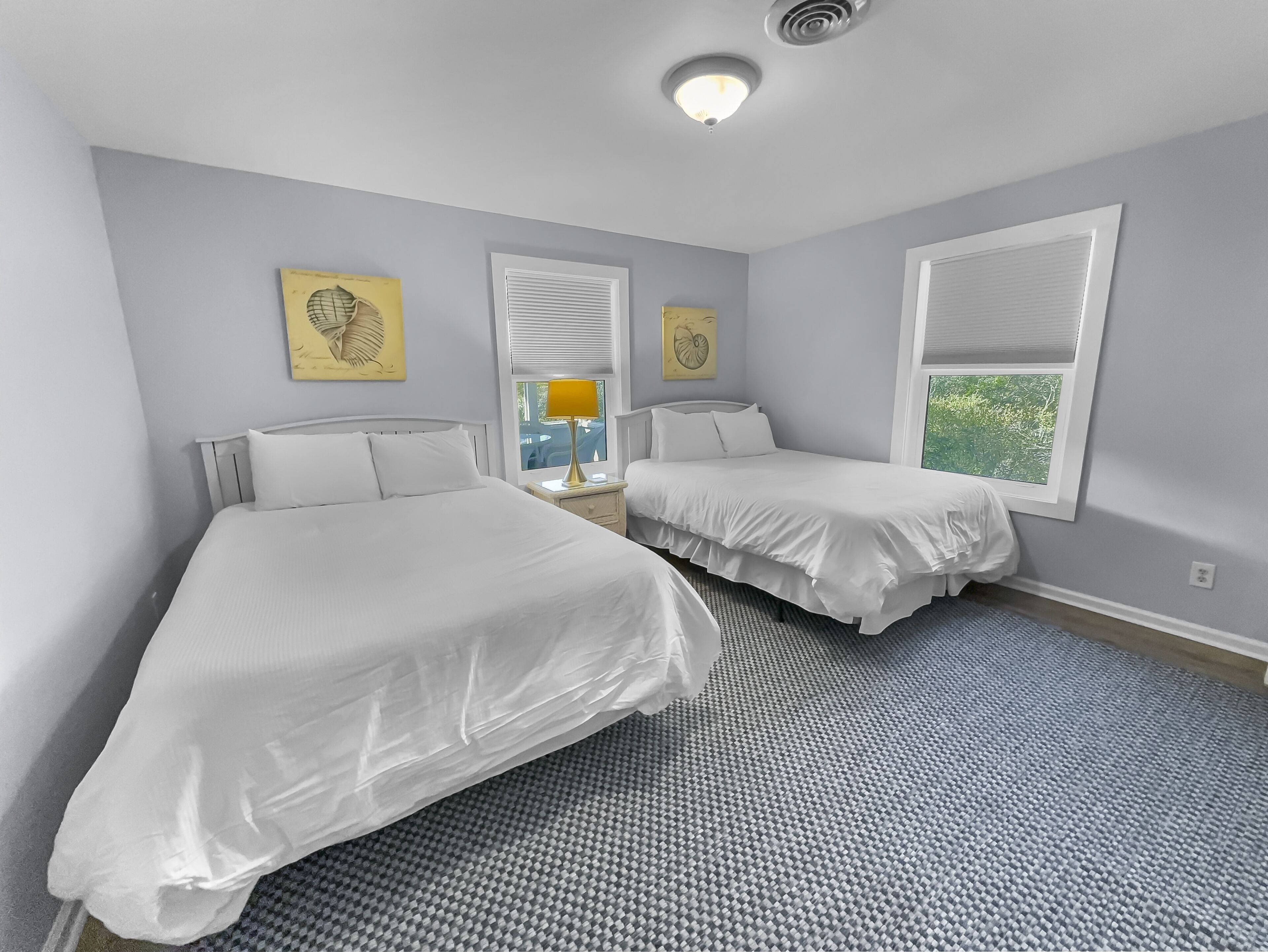 Une chambre avec deux lits simples, une fenêtre avec des stores, un luminaire au plafond et des œuvres d’art accrochées aux murs.