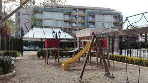 A playground with a yellow slide and swings, surrounded by a fence, with a building in the background. - Sun City Apartments (Sunny Beach)