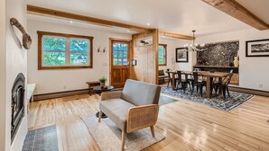 A spacious room with wooden floors, a dining area with a table and chairs, a fireplace, and large windows offering views of greenery.