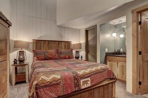 Een slaapkamer met een houten hoofdbord, een gewatteerde spreideken, twee bedlampen, een kleine tafel met een lamp en een spiegel boven de kapstok.