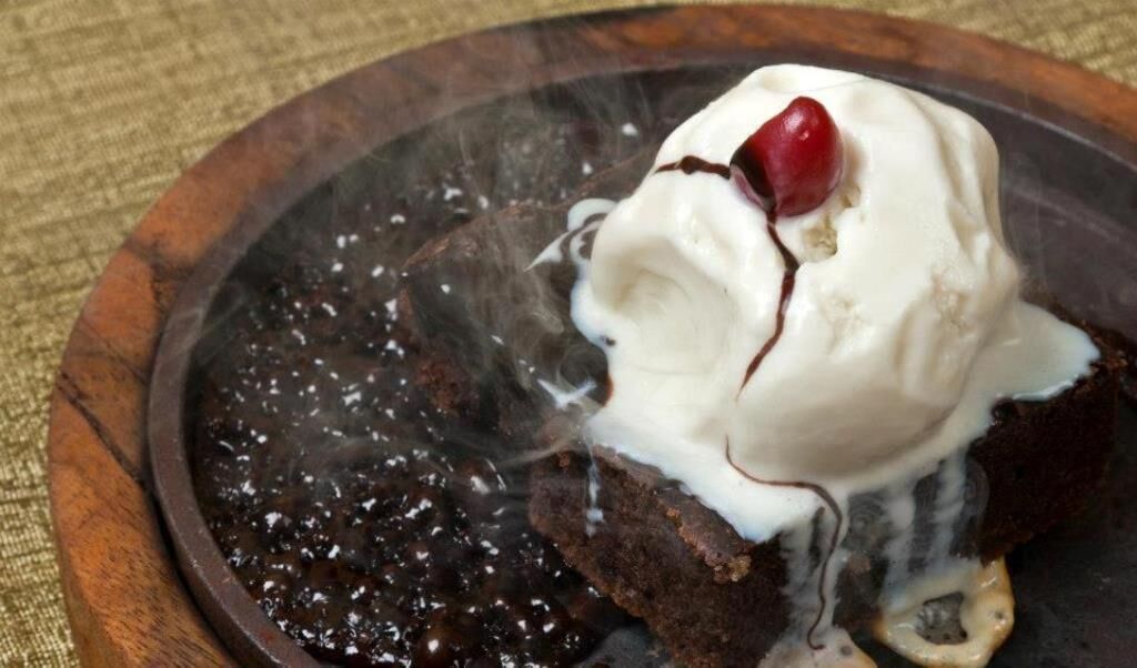 A dessert with whipped cream and a cherry on top, served on a wooden plate.